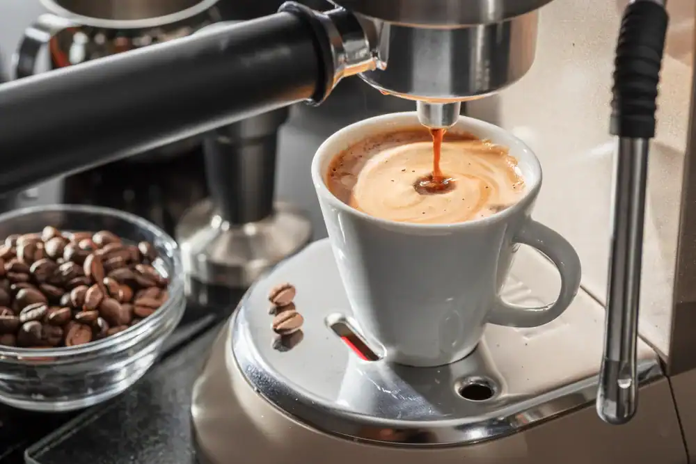 Close-up of an espresso machine pouring coffee into a white cup, with coffee beans in a glass bowl and a few beans on the machine's drip tray.