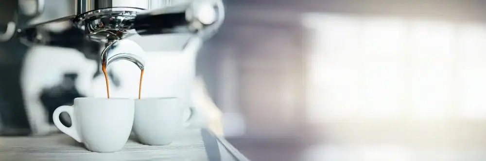 Close-up of an espresso machine pouring coffee into two small white cups, with a blurred background and bright natural light coming from the right side.